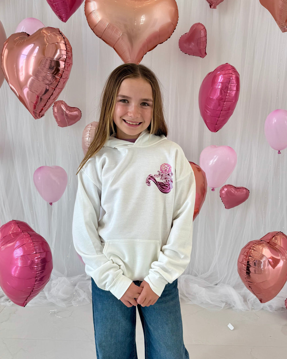 Girl in a white hoodie with a pink Rehna Raiya wave design standing in front of heart-shaped balloons.