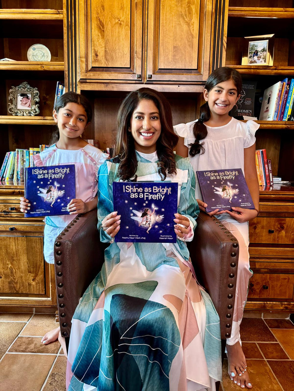 Paperback version of the Children's book titled 'I Shine as Bright as a Firefly' by Neetu Kaur Deol Jhaj with a blue cover featuring a fairy illustration, held by the author and her two children, Rehna and Raiya.