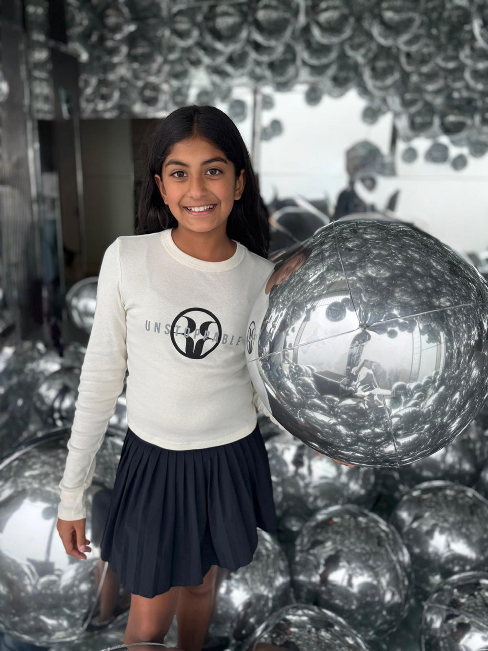 Girl wearing a long sleeve white tee reading "Unstoppable" in silver with the Rehna Raiya logo in black. Paired with a black pleated skirt in a reflective room.