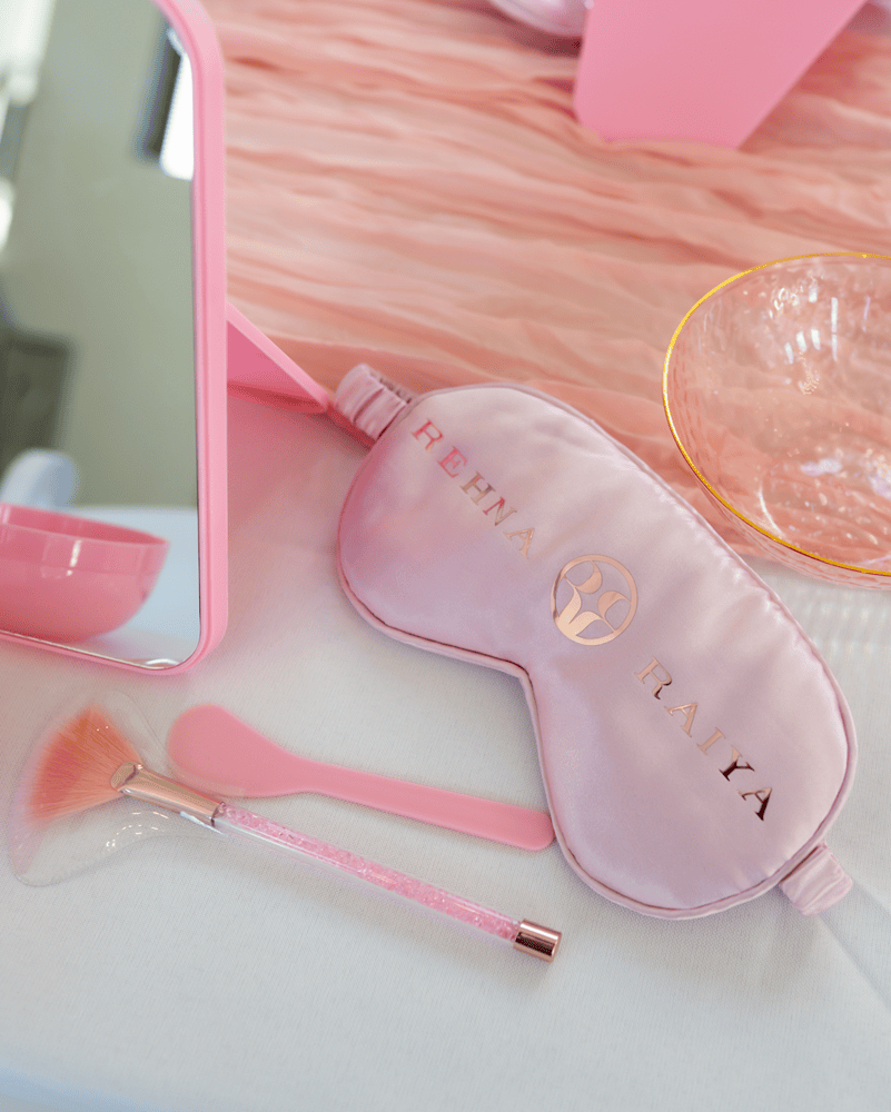 A sleeping mask in pink with rose gold text reading "REHNA RAIYA."